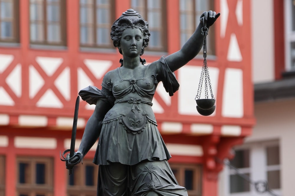Statue de la Justice féminine tenant une balance, devant un bâtiment à colombages rouges et blancs. Automatisation ia pour agence web integré naturellement.